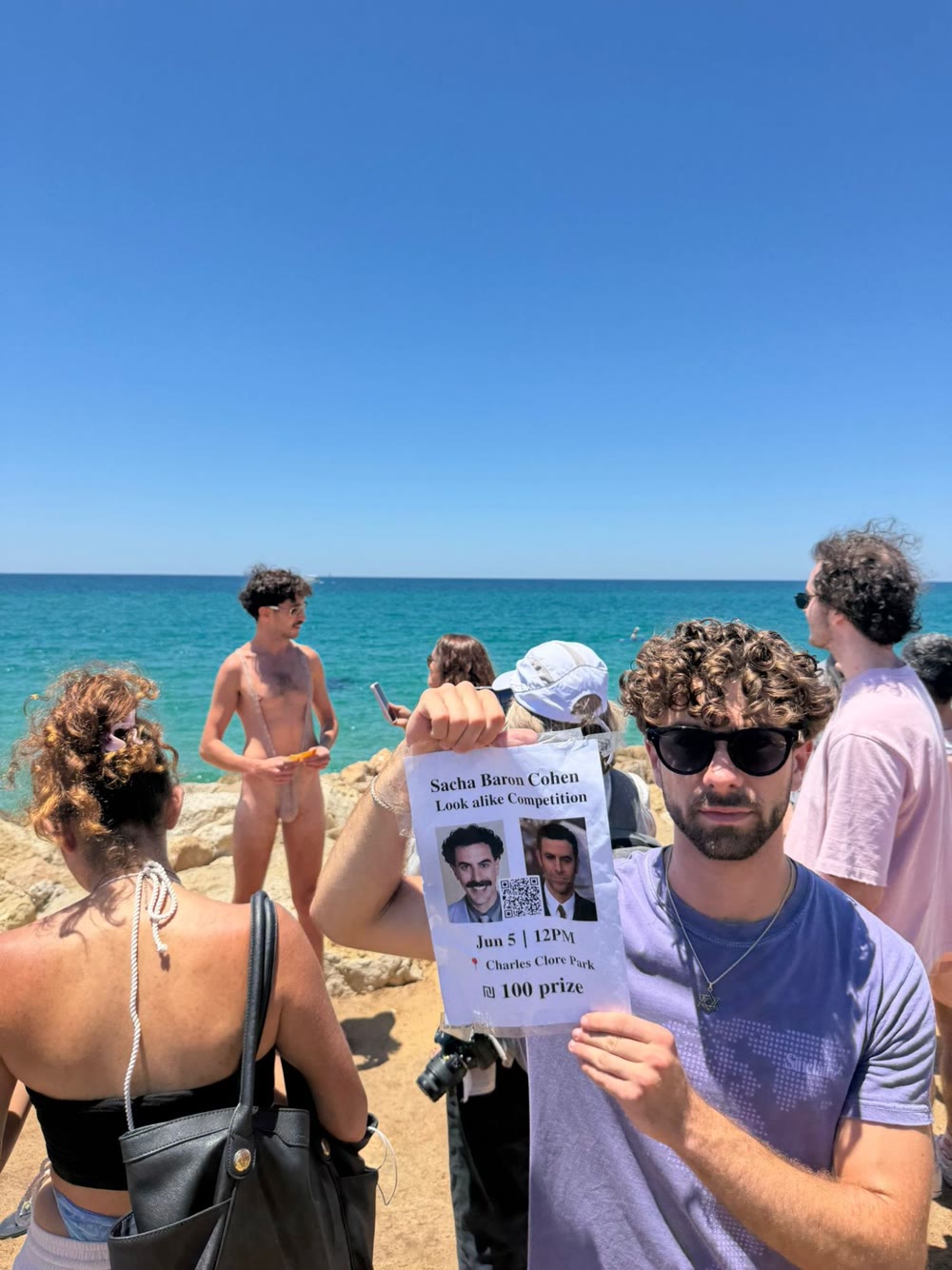 Daniel holding the contest poster on the rocks at Charles Clore Park, with a contestant in a Borat mankini behind him
